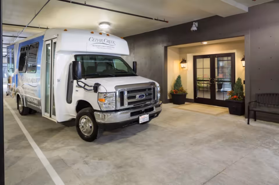 A white and blue shuttle bus parked inside a covered parking area near the entrance of a building. The bus has the text 'Cedar Creek Memory Care Community' on it. The entrance features double glass doors with black frames, flanked by two wall-mounted lantern-style lights and potted plants on either side. A bench is visible to the right of the entrance.