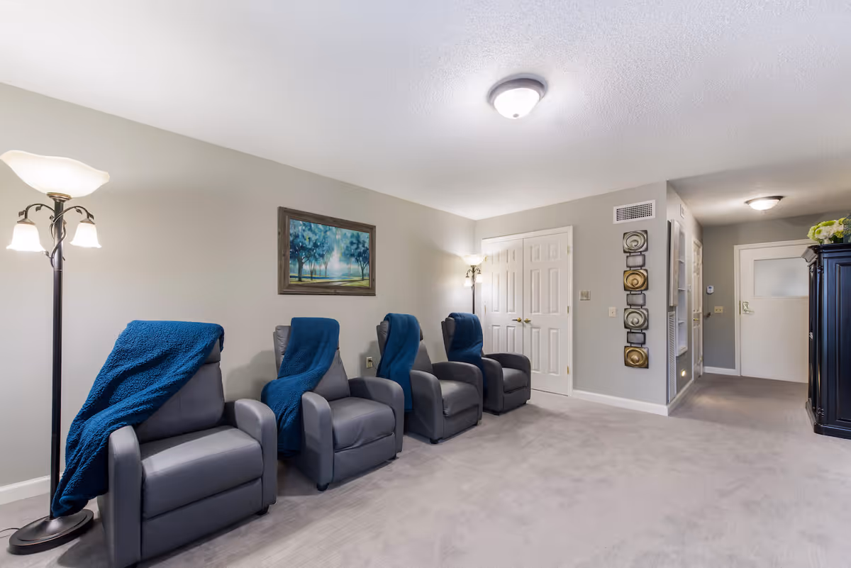 A quiet lounge area with four gray recliner chairs each draped with a blue throw blanket. The room has light gray walls, a carpeted floor, two floor lamps providing warm light, a framed painting of trees on the wall, and decorative wall art near a white double door. A dark wooden cabinet with flowers on top is visible near the hallway entrance.