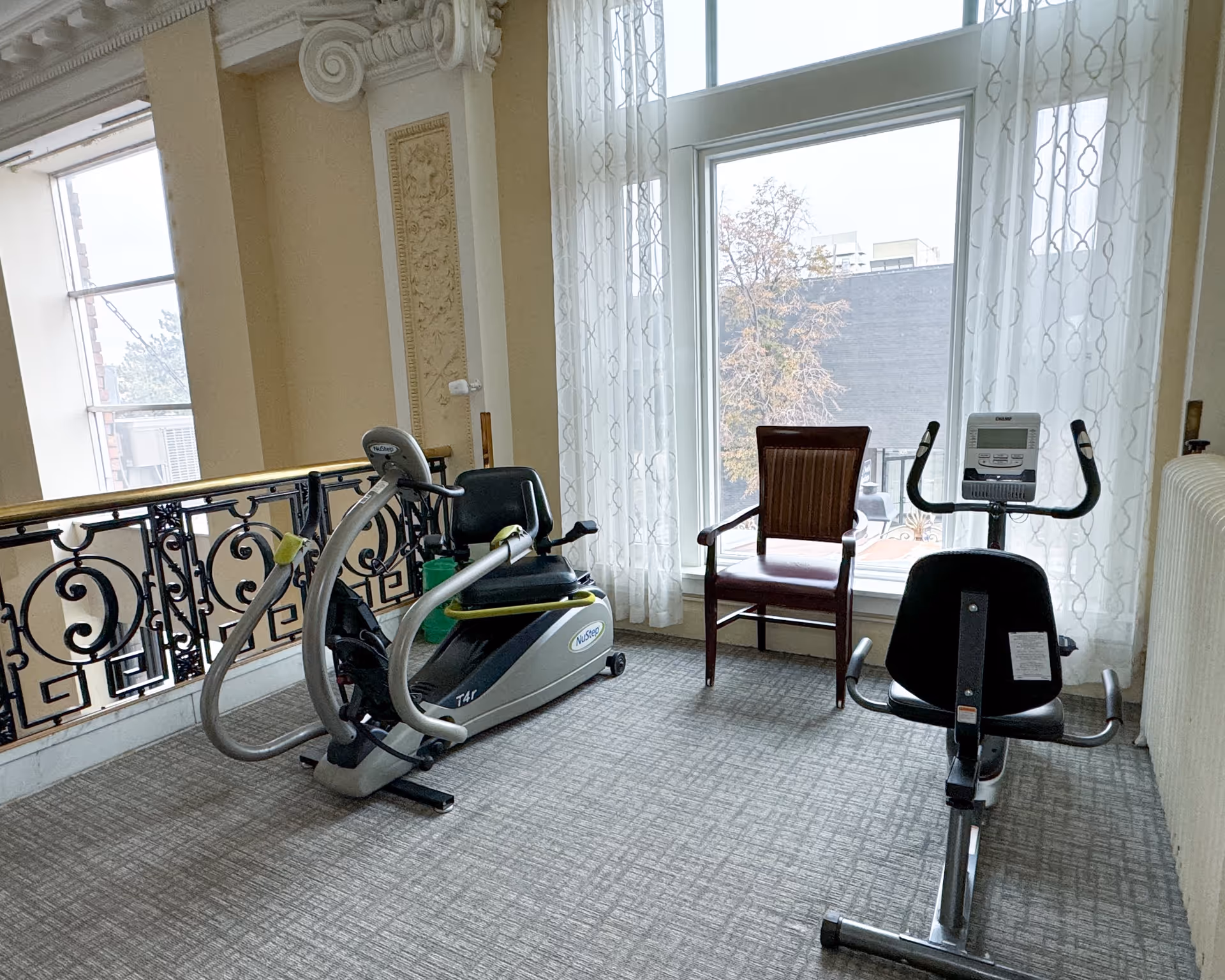 A small exercise area in a senior living facility with two stationary exercise bikes placed on a carpeted floor near large windows with sheer white curtains. There is a wooden chair with armrests positioned between the bikes. The room features ornate molding and a decorative railing along one side.