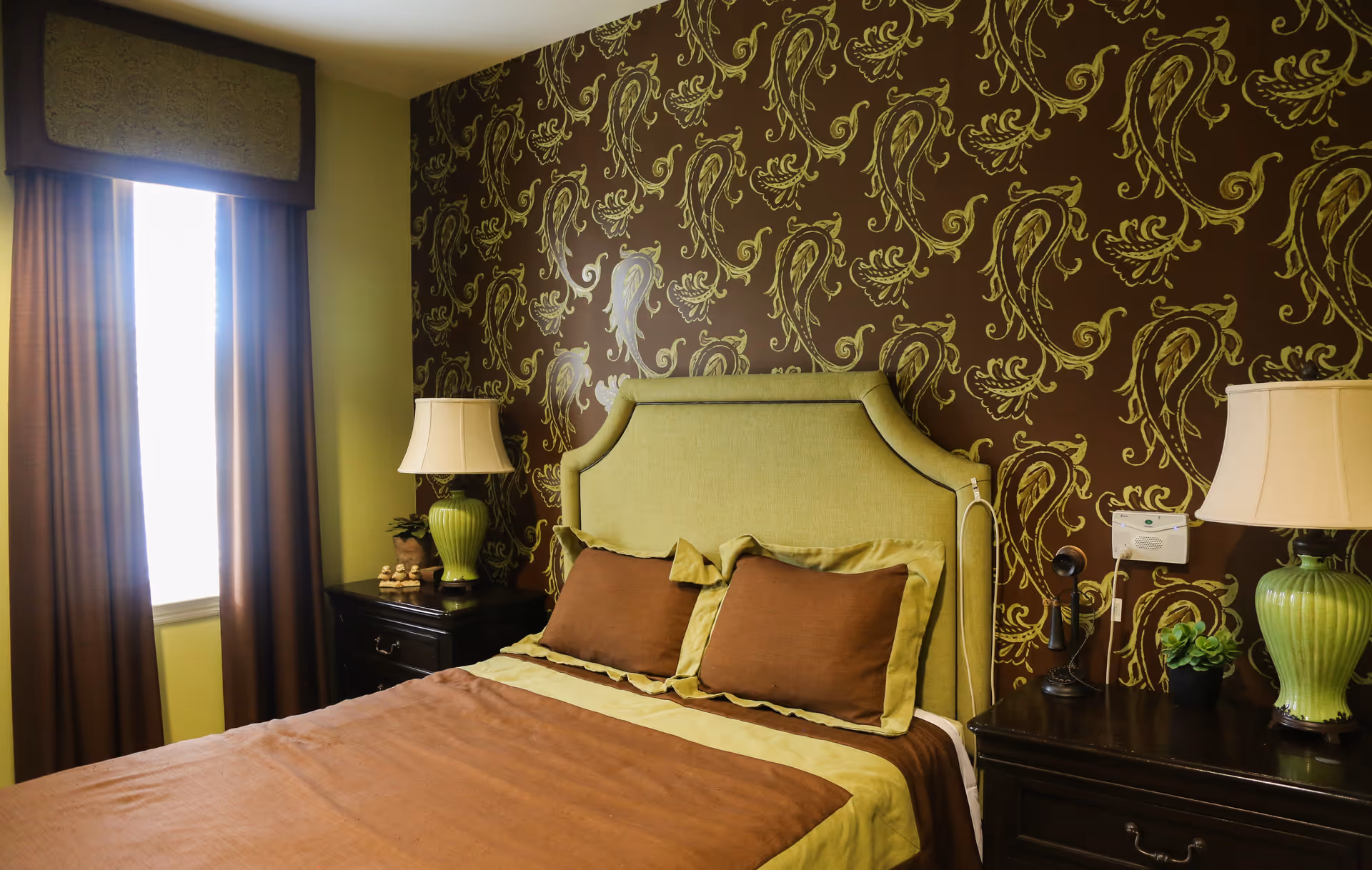 A bedroom with a bed featuring a green upholstered headboard and brown and green bedding. There are two dark wooden nightstands on either side of the bed, each with a green lamp and decorative items. The wall behind the bed has a brown and green paisley pattern wallpaper, and a window with brown curtains is on the left side.