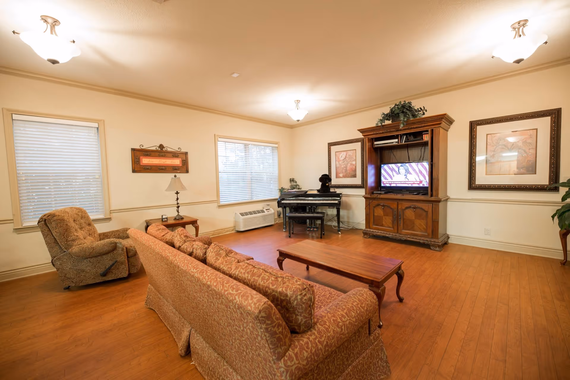 Bright communal living room with patterned sofa and armchair, coffee table, piano, and wooden entertainment center with a TV.