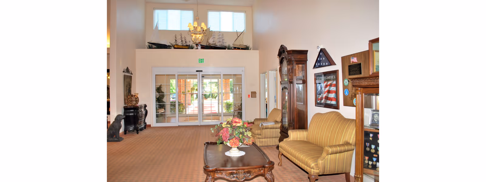 Spacious senior-living lobby with striped sofas and a coffee table topped with flowers, a grandfather clock and display cases facing glass entrance doors.