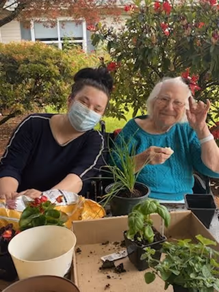 An elderly woman and a younger woman wearing a face mask are sitting outdoors at a table with potted plants and gardening supplies. The elderly woman is smiling and making a hand gesture, while the younger woman is leaning close to her. There are bushes and a building visible in the background.