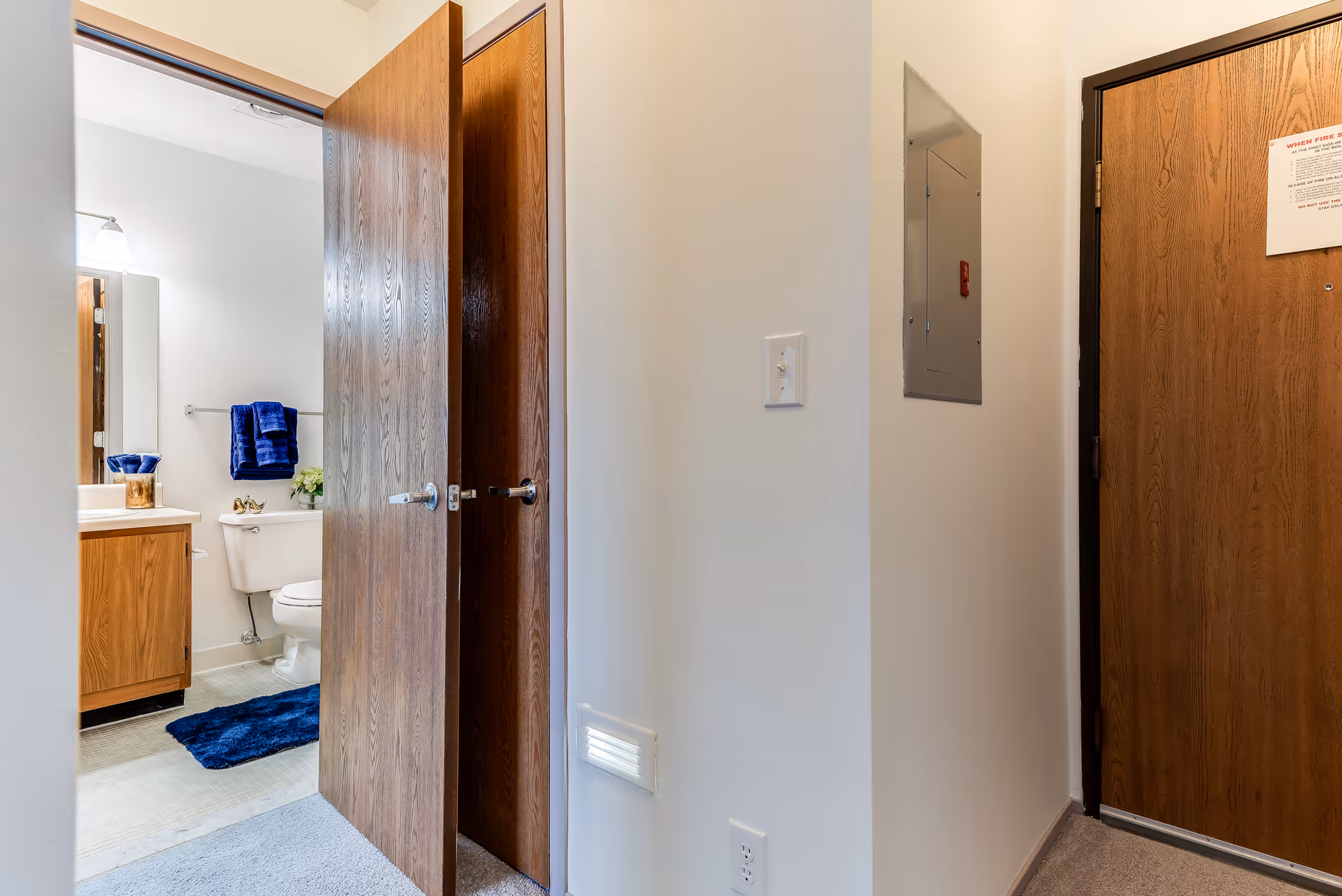 View of a hallway with two wooden doors, one partially open revealing a bathroom with a toilet, sink, mirror, and blue towels and rug. The hallway has light-colored walls, a light switch, an electrical panel, and a closed wooden door with a notice attached.