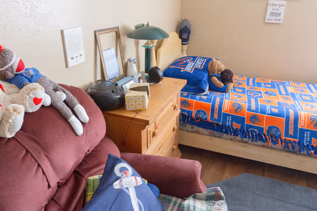A cozy bedroom with a single bed covered in Boise State Broncos themed bedding, including a pillow and blanket. A wooden nightstand beside the bed holds a green lamp, a radio, a tissue box, and some framed pictures. A maroon armchair with stuffed animals and a pillow featuring an anchor design is positioned near the bed. The room has light-colored walls and wooden flooring.