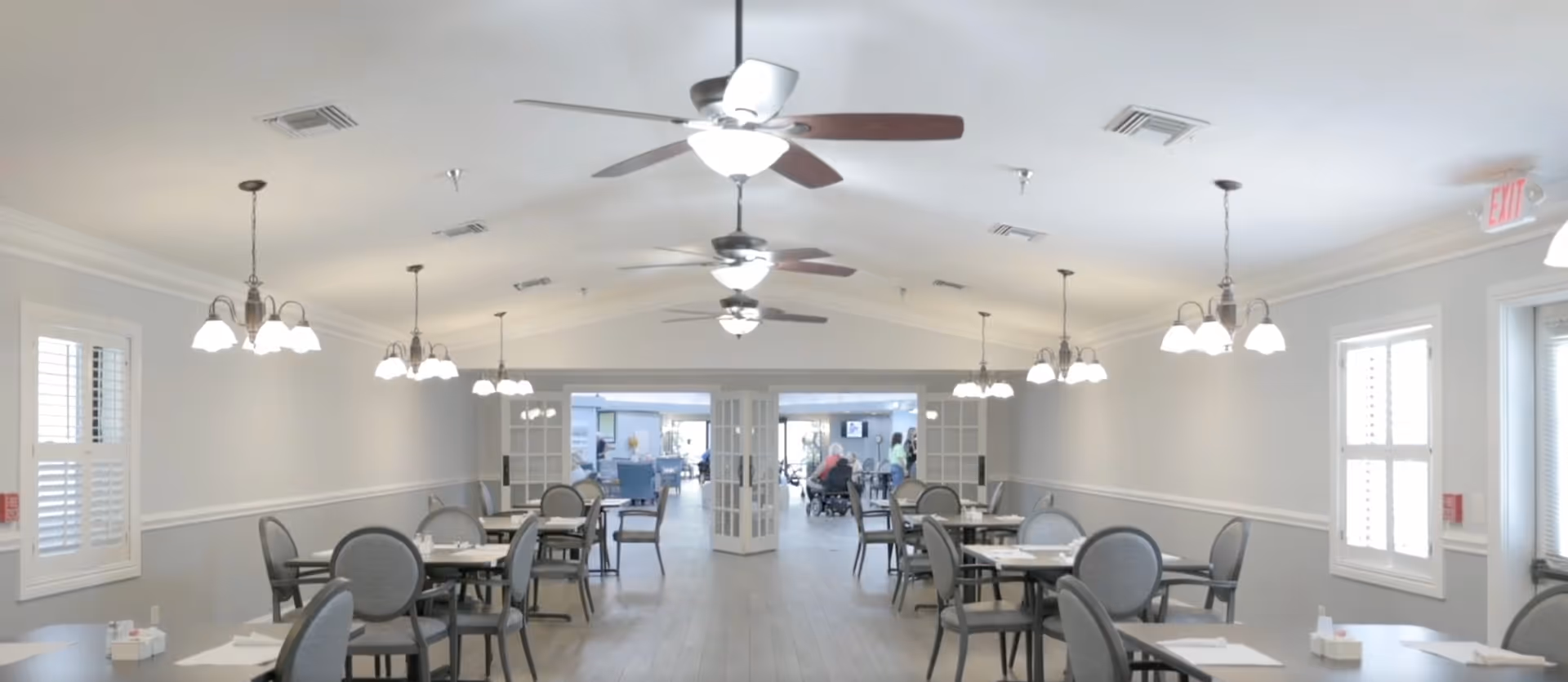 A bright dining room with multiple round tables and gray cushioned chairs arranged neatly. The room has light gray walls, wooden flooring, ceiling fans with lights, and several hanging light fixtures. Large windows with white shutters allow natural light to enter. In the background, there is an open doorway leading to another room where people are visible.