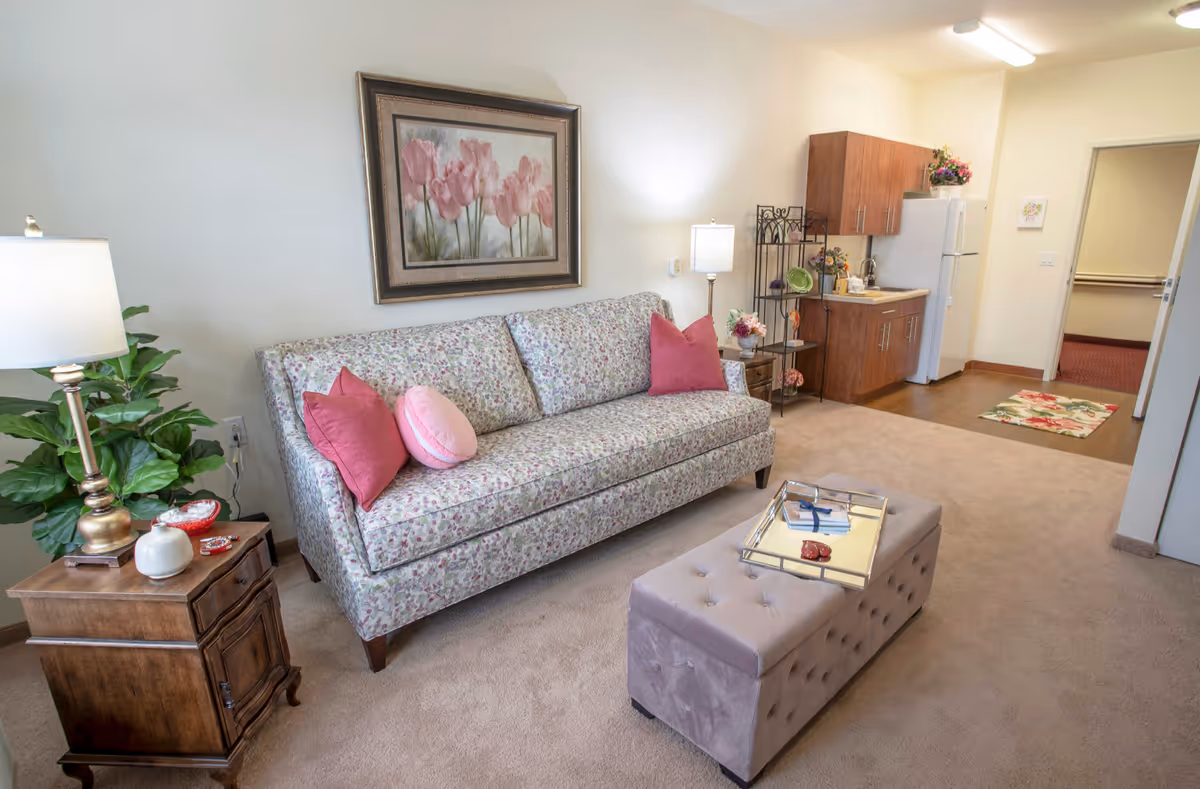 Cozy living room with a floral sofa, pink pillows, tufted ottoman, side table lamp, and a small kitchenette visible in the background.