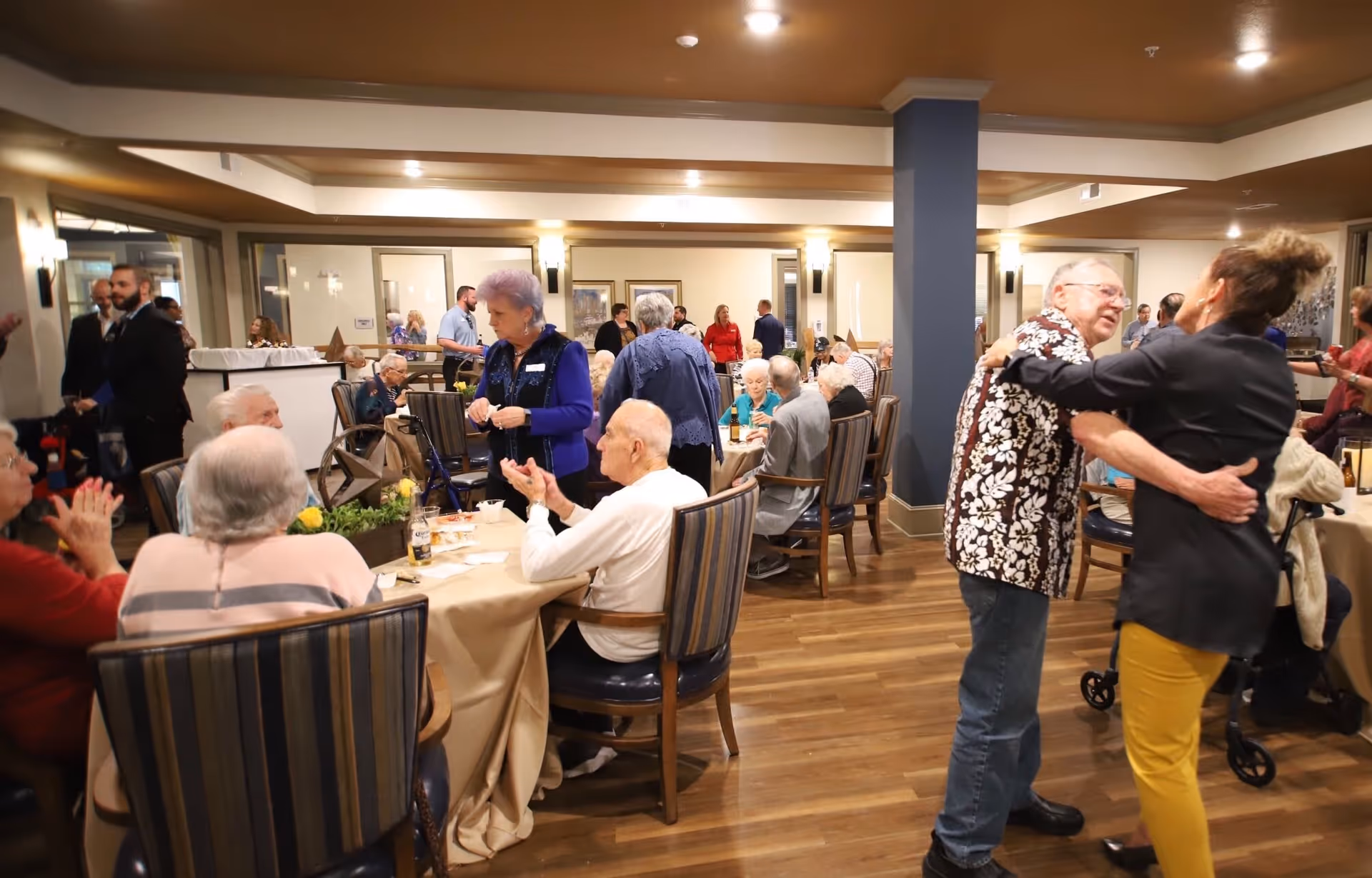 A lively gathering of elderly people socializing and dining in a spacious, well-lit room with wooden floors and comfortable chairs. Some individuals are seated at tables covered with beige tablecloths, while others are standing and engaging in conversation or dancing. The atmosphere appears warm and friendly with a mix of casual and semi-formal attire.