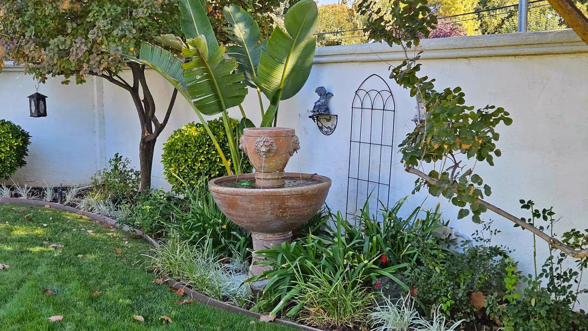 A garden area with a stone water fountain surrounded by various green plants and bushes. There is a white wall in the background with a decorative metal trellis and a small wall-mounted sculpture. The grass is neatly trimmed and there are some fallen leaves scattered on it.