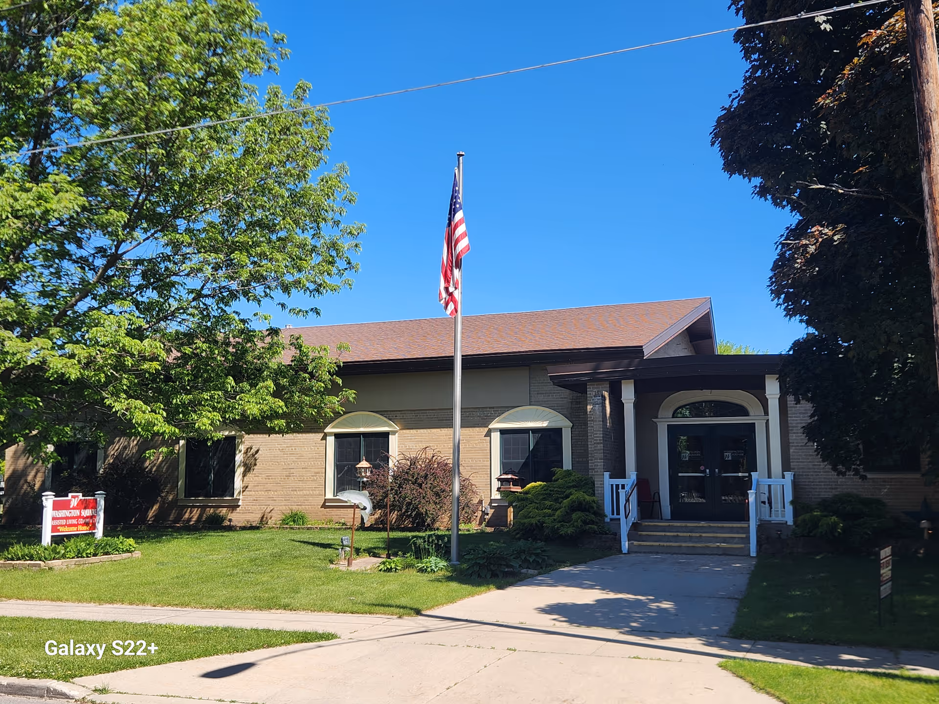 Front exterior of Washington Square Assisted Living building with an American flag, lawn, and main entrance walkway.