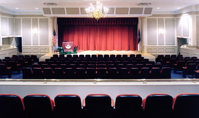 An auditorium with rows of red cushioned seats facing a stage with a closed red curtain. The stage has an American flag on the left, a podium with a sign, and a chandelier hanging from the ceiling. The walls are decorated with white paneling and there are doors on either side of the stage.