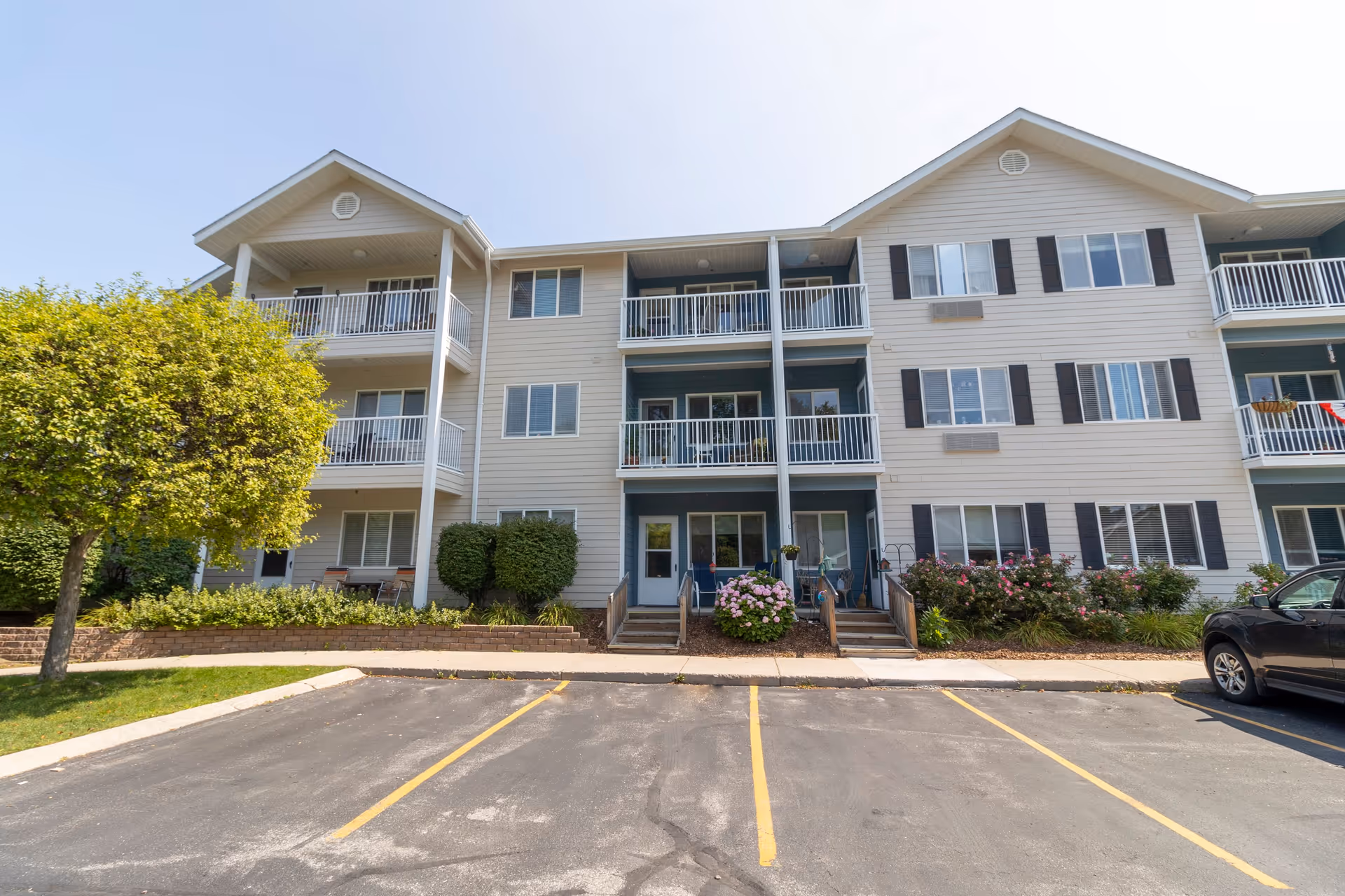 Exterior view of a three-story residential building with balconies, windows, and a small landscaped area with bushes and a tree in front. There is a parking lot with marked spaces in the foreground under a clear blue sky.