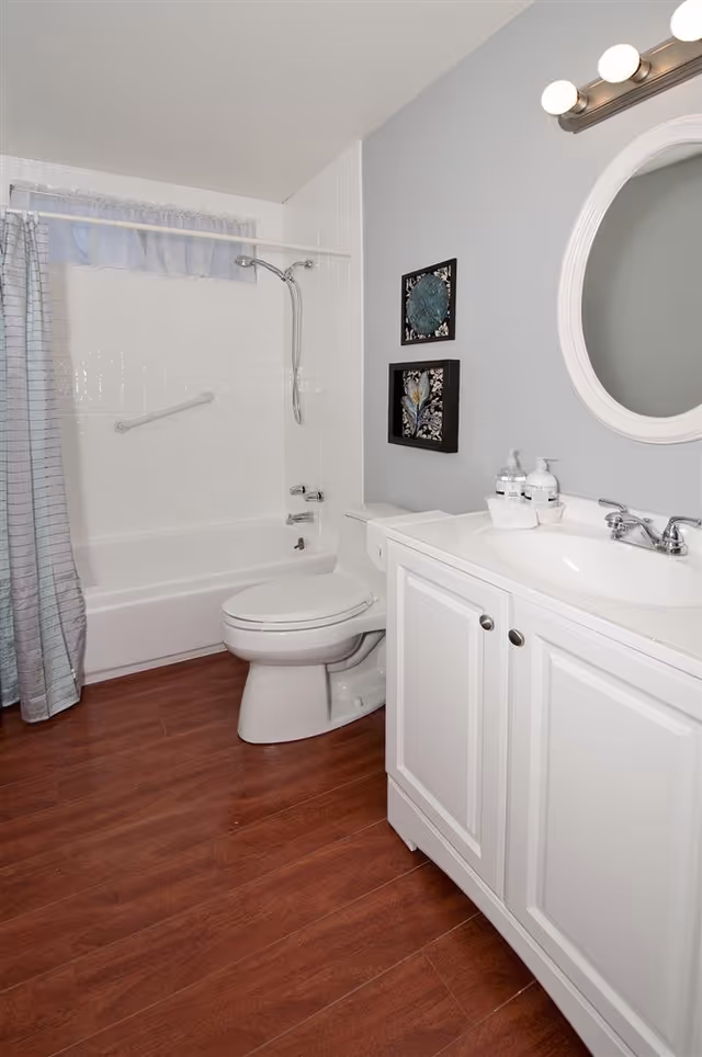 A clean bathroom with a white bathtub and shower combination, a white toilet, and a white vanity with a sink and mirror above it. The walls are painted light gray, and there are two framed decorative pieces hanging on the wall. The floor has wood-like brown tiles, and a shower curtain is partially visible.