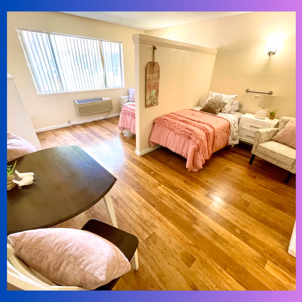 Bright shared bedroom with two twin beds separated by a half wall, hardwood floors, a small table and chairs, and a window with vertical blinds.