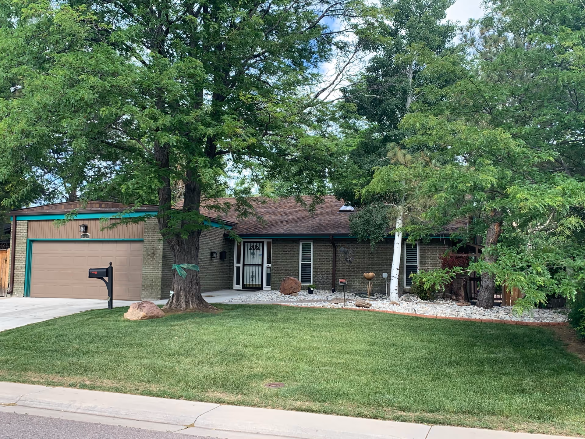 Single-story brick house with a brown roof and a two-car garage. The front yard has a well-maintained green lawn, several large trees, and decorative rocks near the entrance. A black mailbox is positioned near the driveway.