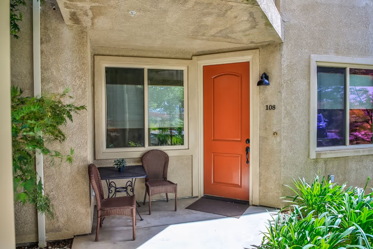 Outdoor entrance area of a residential unit with a red door numbered 108, two brown wicker chairs, a small black table with a plant on it, and green plants along the side.