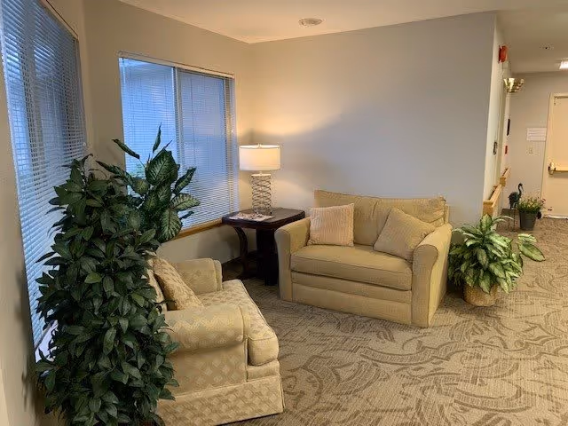 A cozy interior seating area with two beige upholstered armchairs and a loveseat arranged around a small wooden side table with a lamp. There are two large potted plants near the seating area, and windows with blinds on the left wall. The carpet has a patterned design, and the walls are painted light beige.