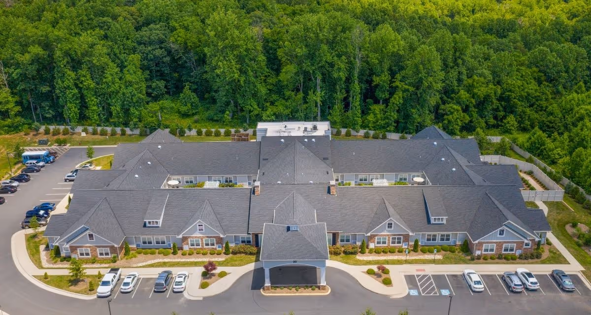 Aerial view of a single-story memory care building with parking lots in front and a wooded area behind.