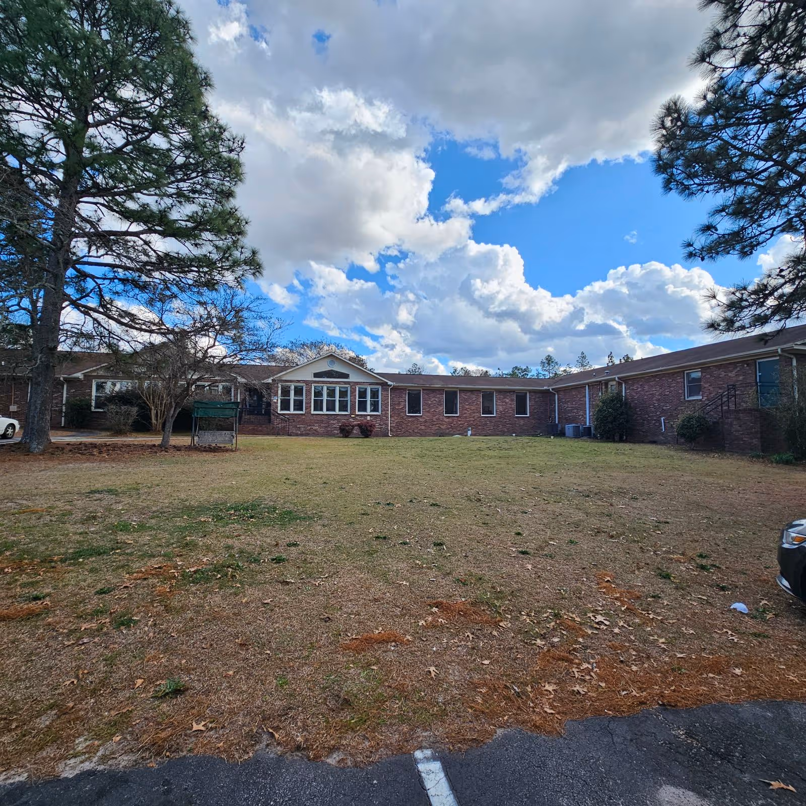 Exterior view of a single-story brick building with multiple windows, surrounded by a grassy lawn and trees under a partly cloudy blue sky. A car is partially visible on the right side of the image.
