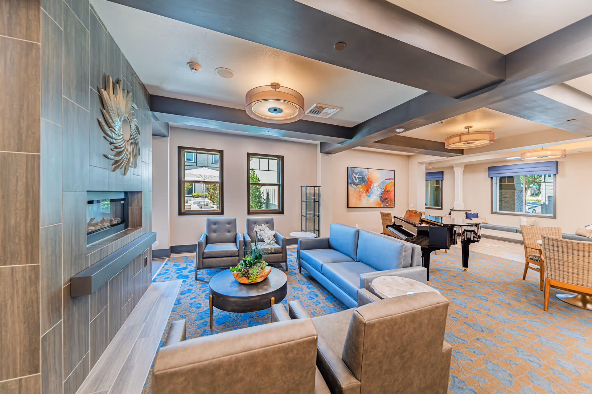 A spacious and well-lit senior living facility common area featuring a modern fireplace with a decorative sunburst mirror above it, a blue sofa, multiple armchairs, a round coffee table with a plant centerpiece, a grand piano, and a dining area with wicker chairs. The room has large windows, ceiling beams, and contemporary light fixtures.