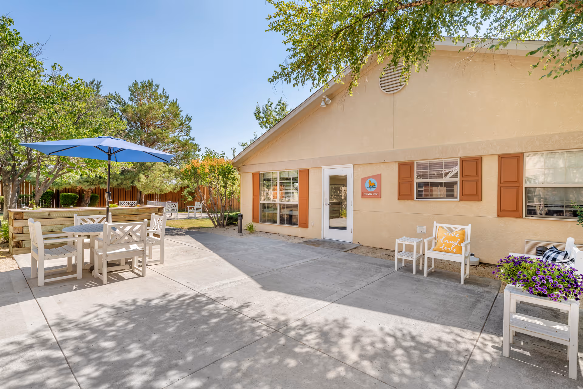 Sunlit outdoor patio with white tables and chairs, a blue umbrella, and a beige building with shuttered windows.