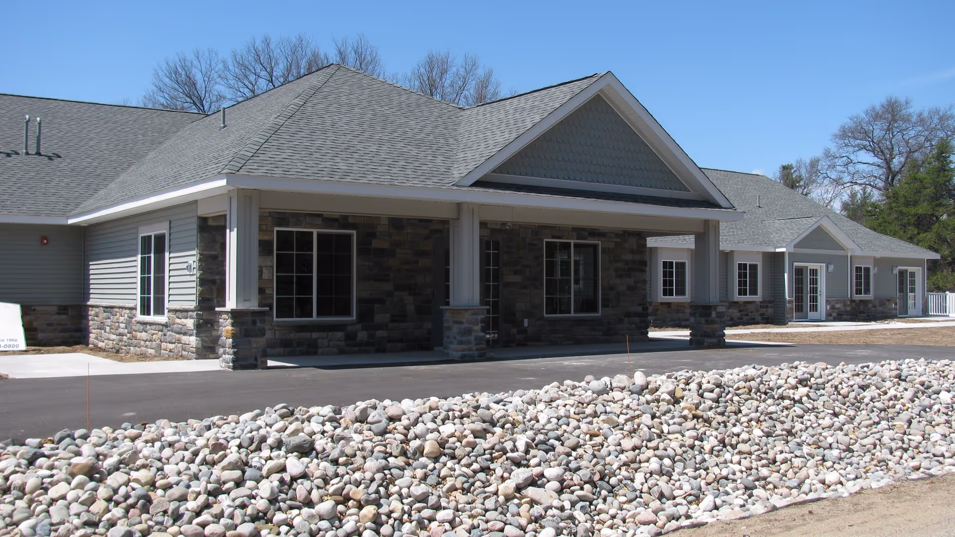 Exterior view of a single-story assisted living facility building with a covered entrance supported by columns. The building features stone and siding exterior walls, multiple windows, and a paved driveway. In the foreground, there is a landscaped area filled with small rocks. Trees are visible in the background under a clear blue sky.