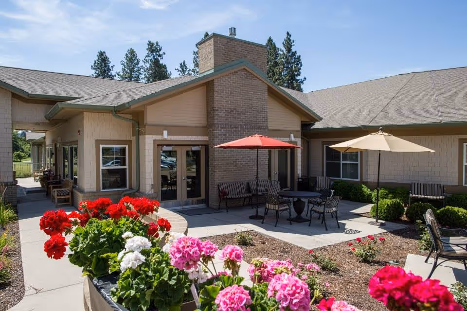 Outdoor patio area at Orchard Ridge Senior Living featuring a building with beige siding and a brick chimney. The patio has tables with umbrellas, chairs, benches, and landscaped flower beds with red, pink, and white flowers under a clear blue sky.