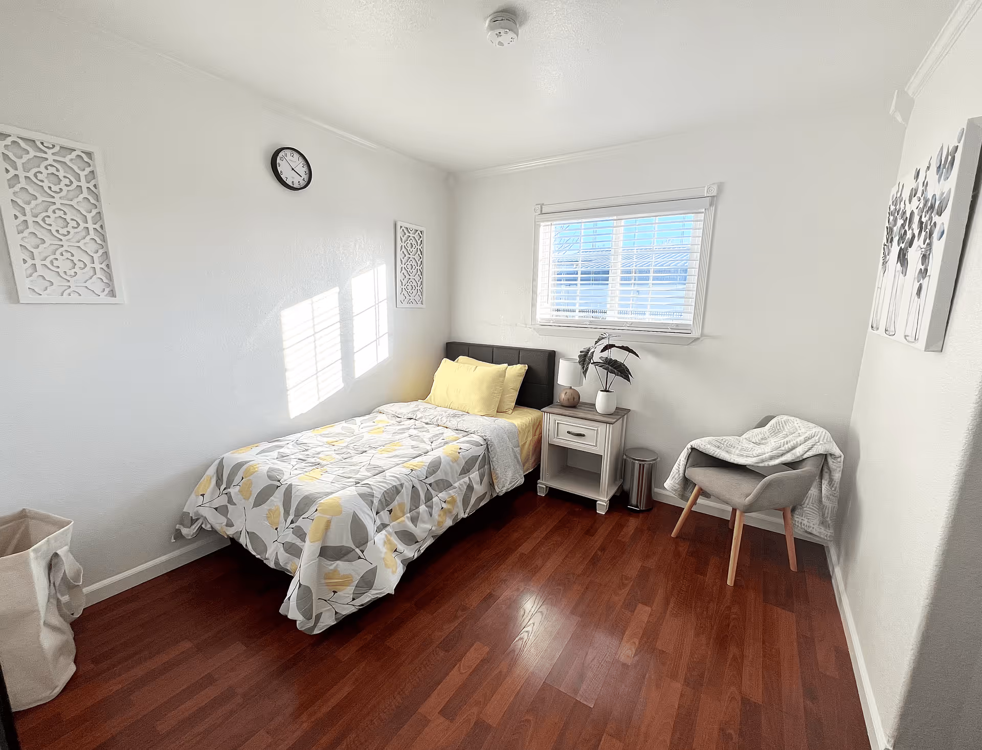 Small, bright bedroom with a single bed, nightstand, chair, and a window letting in sunlight.