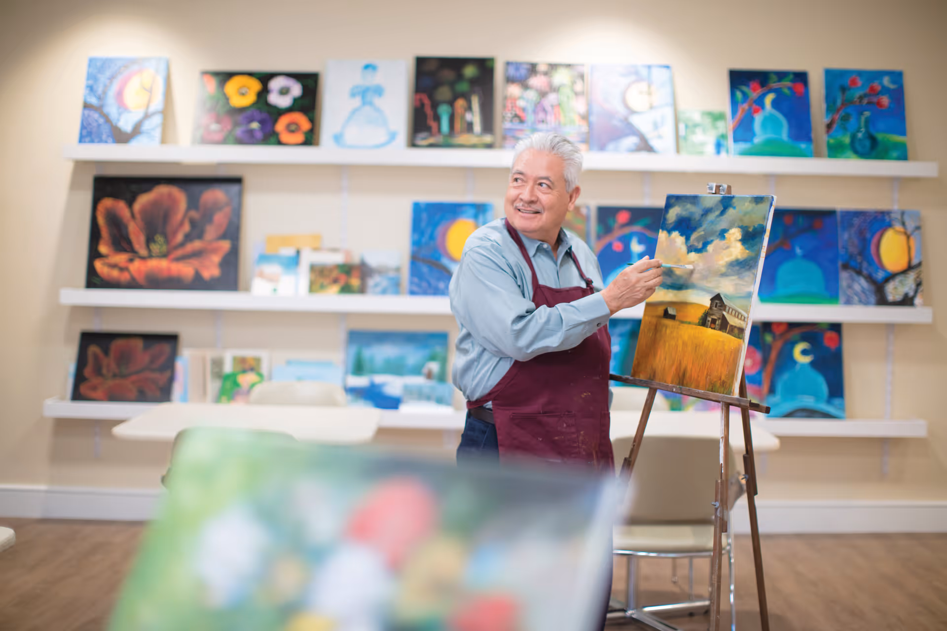An elderly man wearing a maroon apron is painting a landscape on an easel in an art room. Behind him, there are shelves filled with various colorful paintings. The room has a warm and inviting atmosphere.