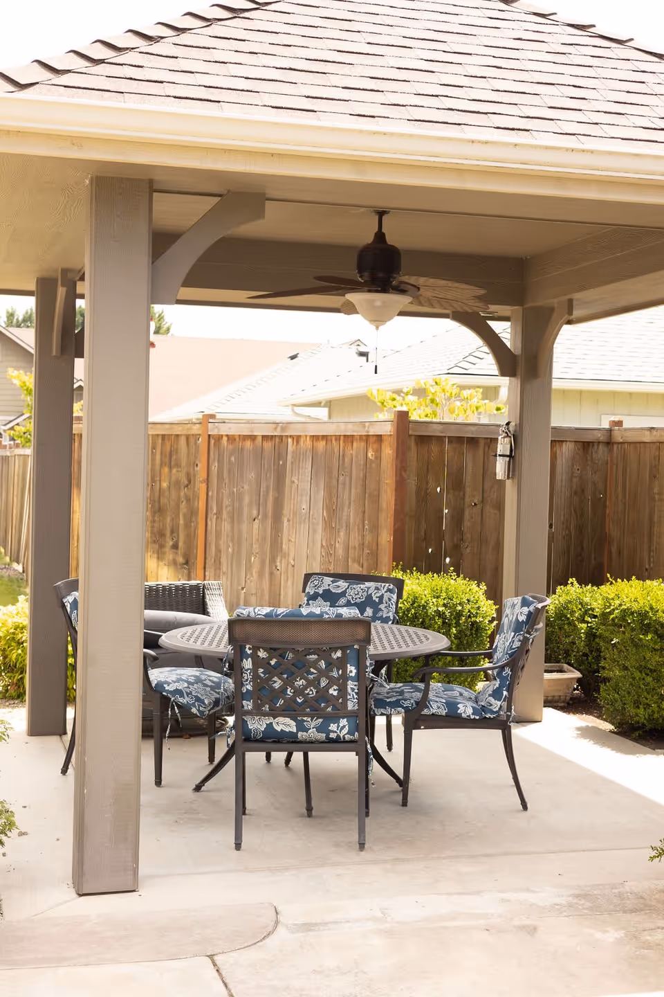 Outdoor covered patio area with a round metal table and five cushioned chairs with blue floral patterns. The patio has a ceiling fan with a light fixture and is surrounded by a wooden fence and green bushes.