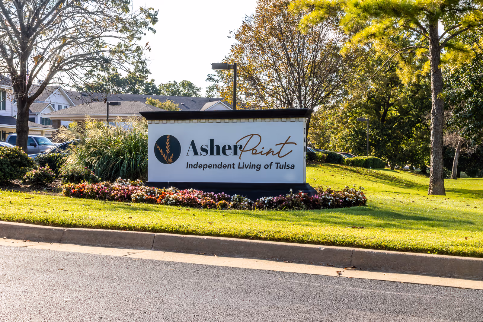 Outdoor view of a sign for Asher Point Independent Living of Tulsa surrounded by green grass, flowers, trees, and a paved road in front.