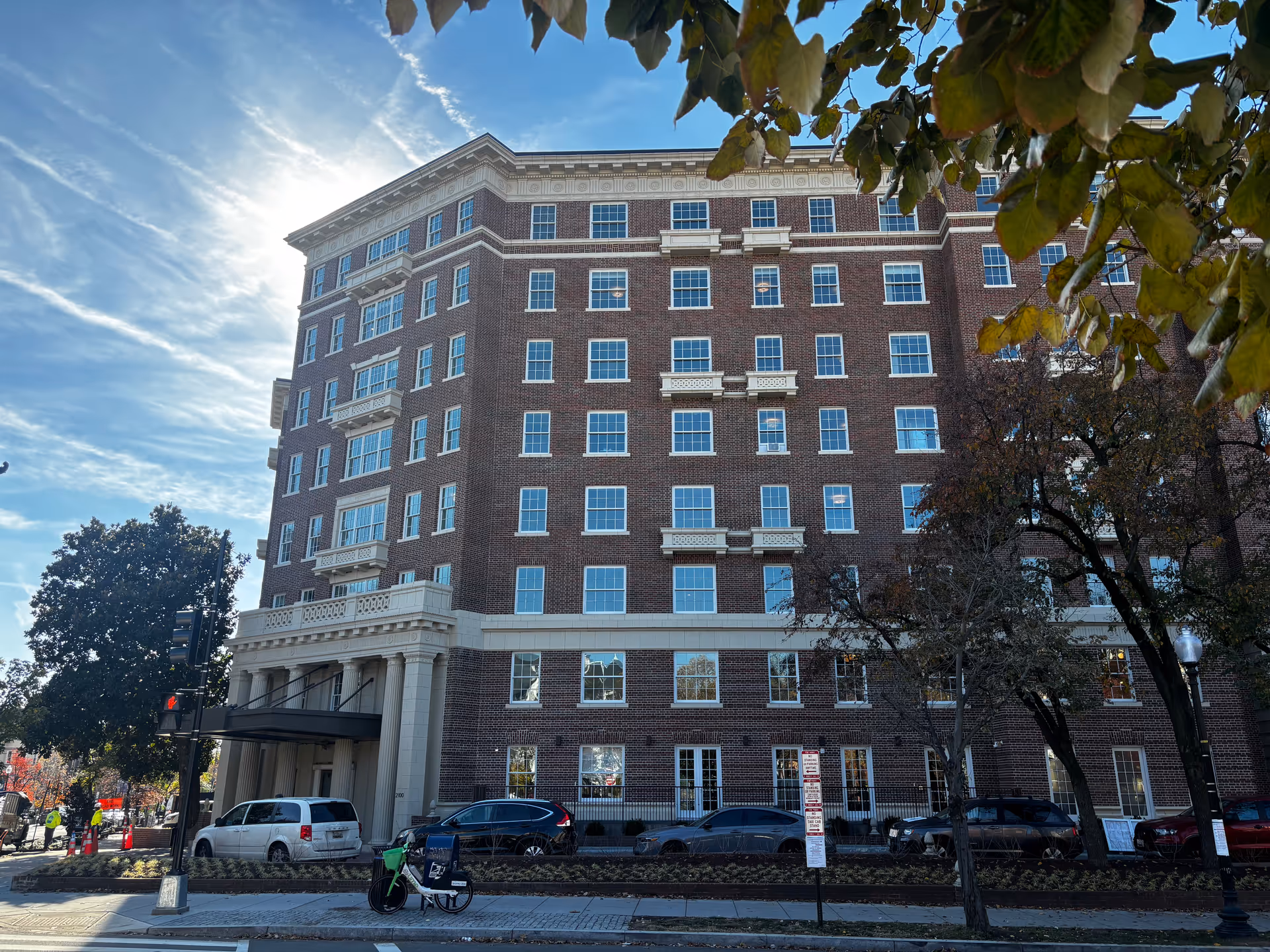 Front facade of a multi-story brick building with parked cars and trees along the street.