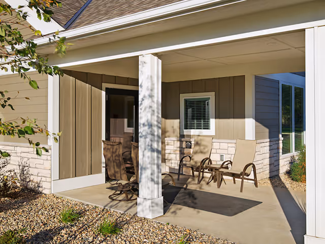 Covered outdoor patio area with four beige cushioned chairs and a small table on a concrete floor, surrounded by a gravel garden bed with some plants. The patio is attached to a building with beige siding and stone accents, and there is a white column supporting the roof.