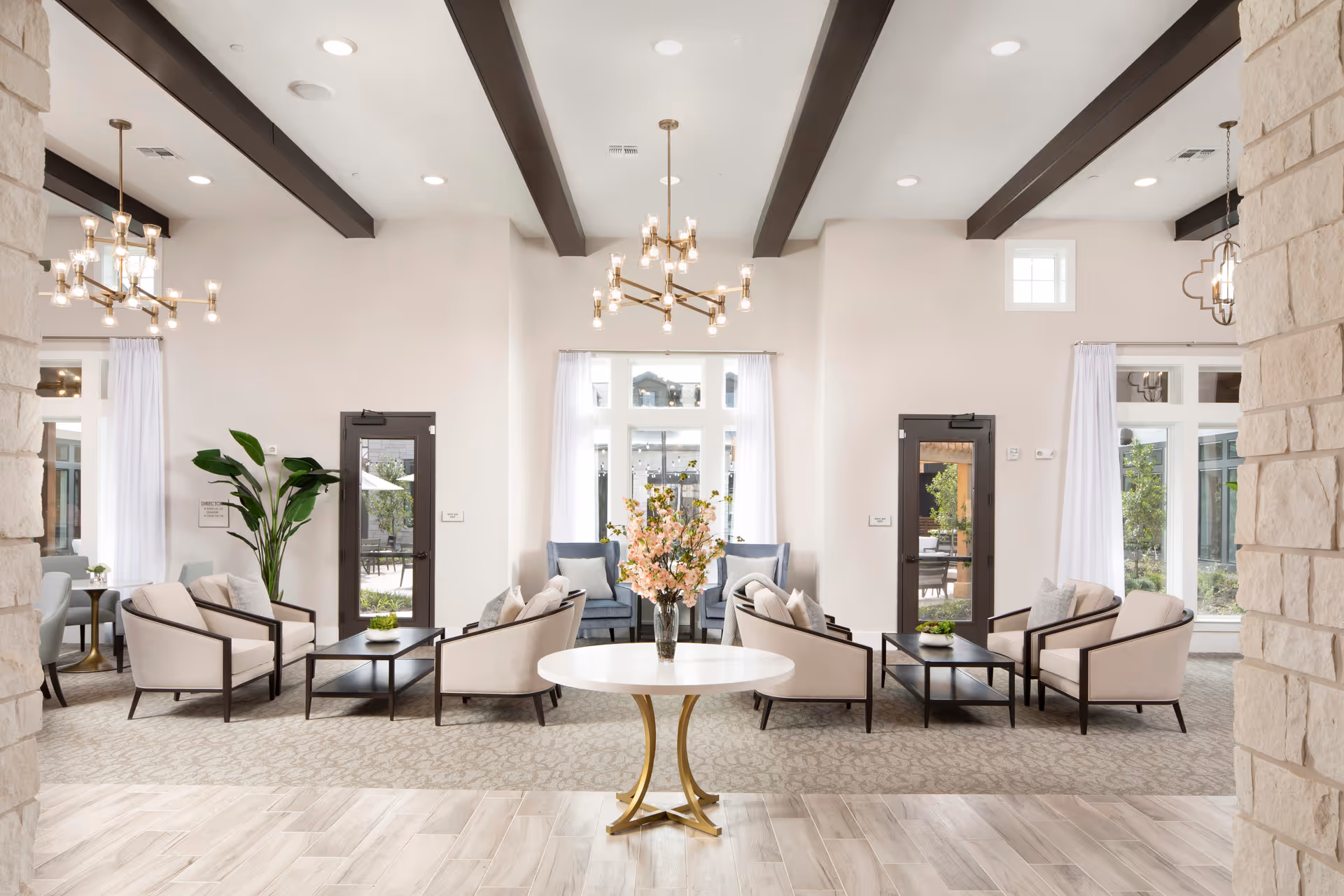 Bright, spacious senior living lounge with multiple seating areas, chandeliers, and a central round table topped with flowers.