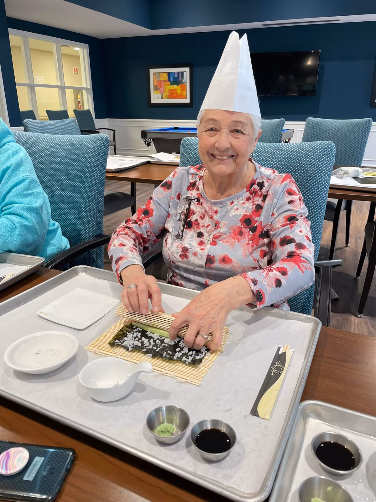 An elderly woman wearing a white chef's hat and a floral shirt is sitting at a table in a dining area, smiling while rolling sushi on a bamboo mat. There are small bowls with soy sauce and wasabi, chopsticks, and other sushi-making items on the tray in front of her. The room has blue walls, teal chairs, and a pool table in the background.