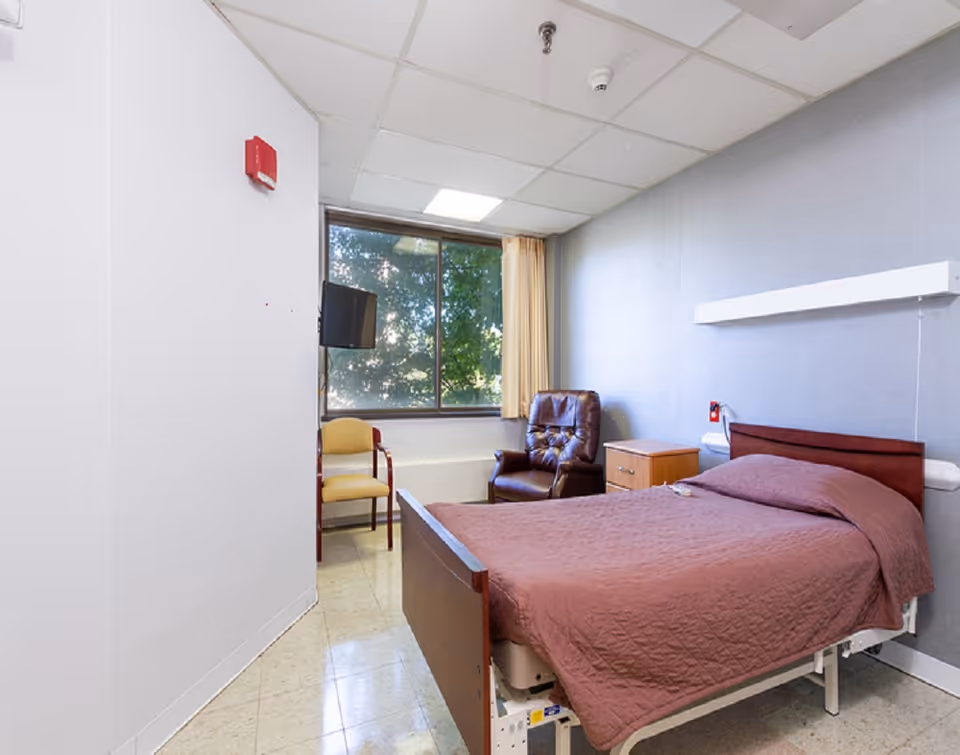 A small senior care patient room with a single adjustable bed, bedside table, leather recliner and a window with curtains.