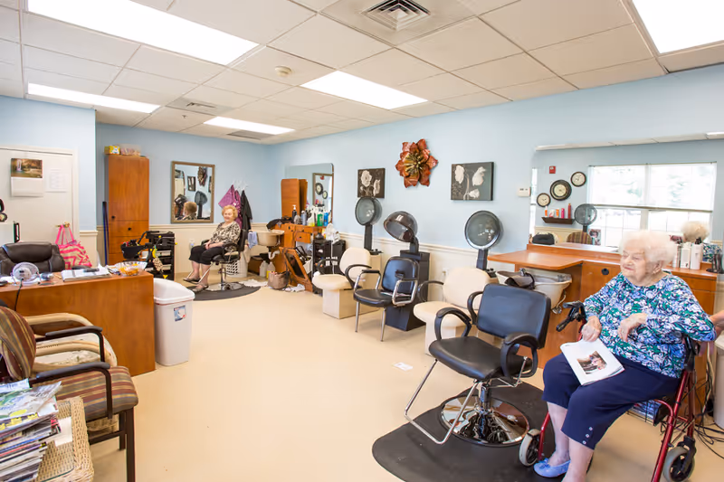 Interior of a senior living facility hair salon with two elderly women seated. The room has light blue walls, salon chairs, hair dryers, mirrors, and various hair care products on counters. One woman is sitting in a salon chair near the back, and another woman is seated in a wheelchair holding a magazine near the front.