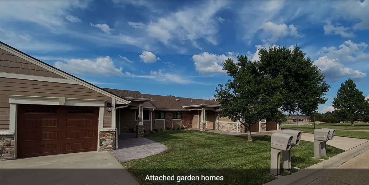 Attached garden homes with brown garage doors, beige siding, stone accents, a green lawn, trees, and a blue sky with scattered clouds.