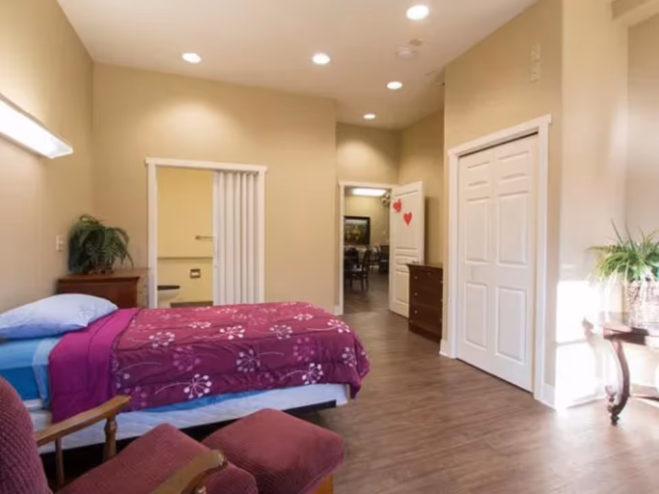 Well-lit bedroom with a single bed covered in a purple floral blanket, an armchair, plants, and a doorway leading to a hallway.