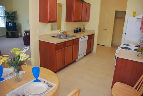 A kitchen area with wooden cabinets, a sink, dishwasher, stove, and refrigerator. A small round dining table with two place settings and blue glasses is visible in the foreground. The kitchen opens into a living room area with a television and a plant near the window.
