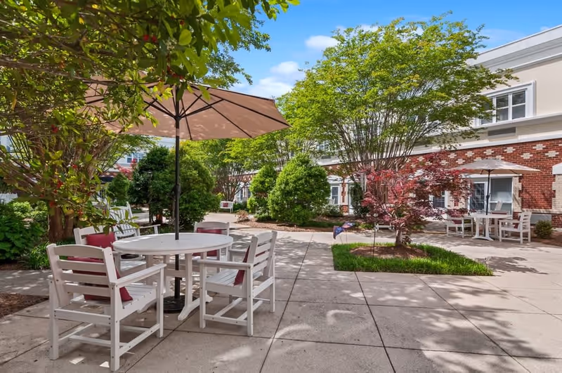 Outdoor courtyard area with white patio tables and chairs, some with red cushions, shaded by large umbrellas. The courtyard is surrounded by green trees and bushes, with a brick and white building in the background under a blue sky with a few clouds.