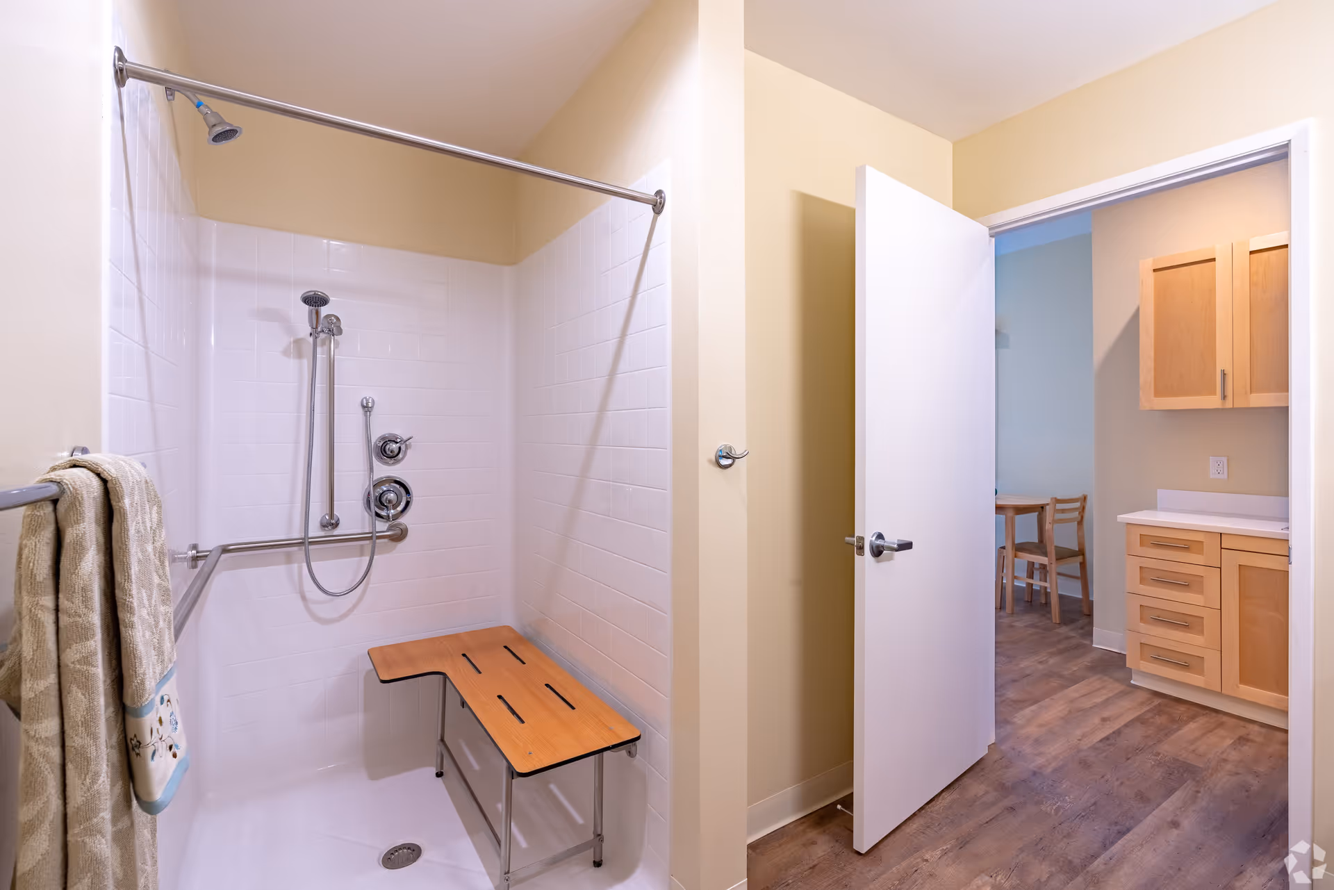 Accessible bathroom shower with grab bars and a wooden fold-down bench, with an open door showing a kitchen/dining area.