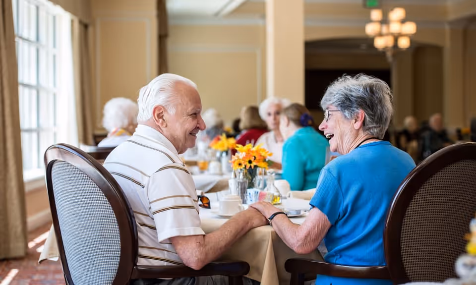 An elderly man and woman sitting at a dining table in a well-lit room, holding hands and smiling at each other. Other elderly people are seated at tables in the background, with flowers and dining items on the tables.