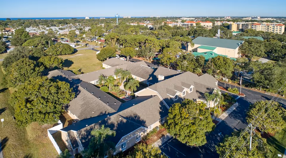 Aerial view of Maris Pointe senior living facility showing multiple connected buildings with gray roofs surrounded by trees and greenery. The facility is located in a suburban area with other buildings and a water tower visible in the background under a clear blue sky.