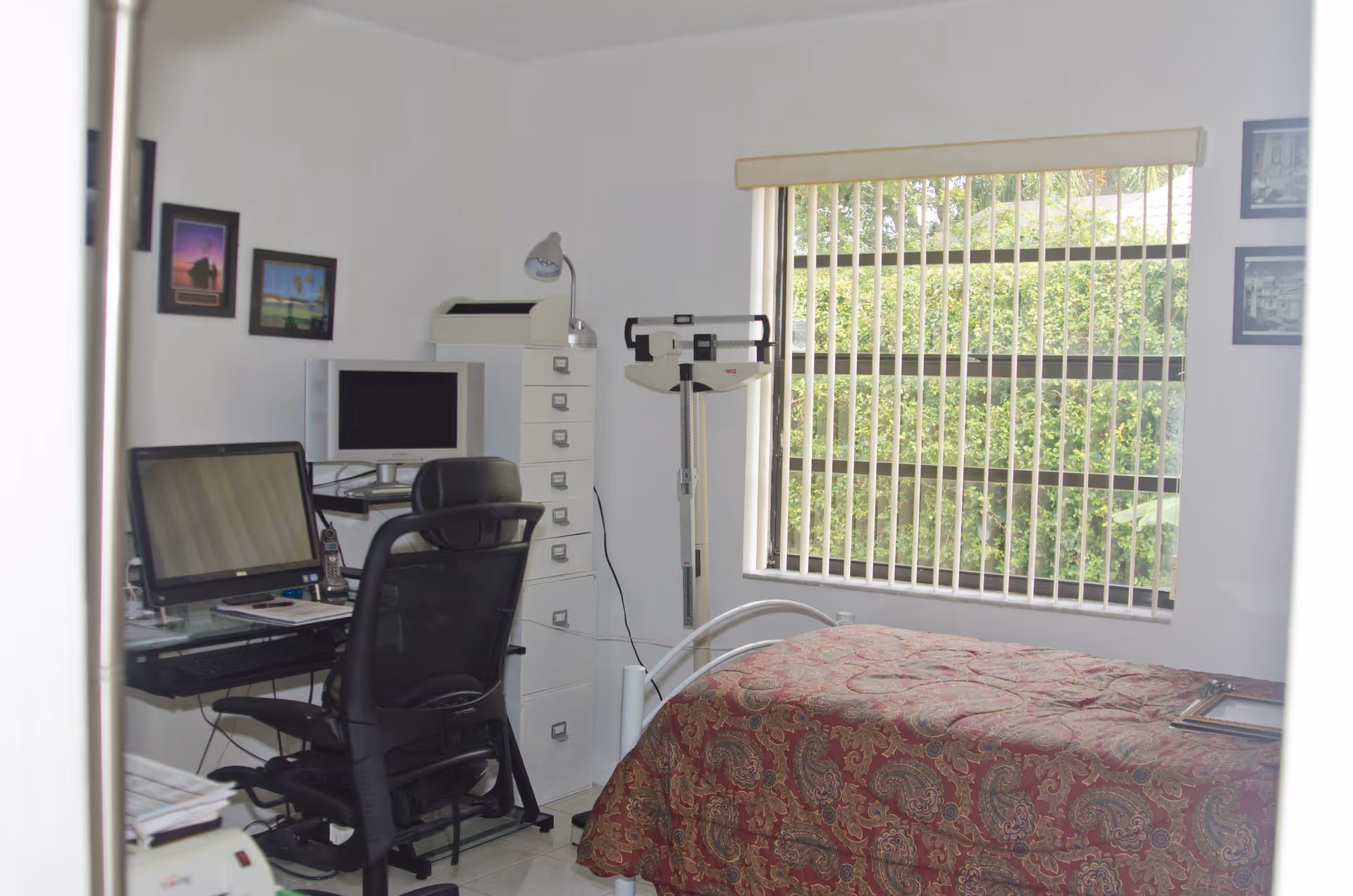 A small room with a single bed covered with a patterned red and gold bedspread. Next to the bed is a window with vertical blinds showing greenery outside. There is a black office chair and a desk with two computer monitors, a phone, and some papers. A tall white filing cabinet with a desk lamp on top stands beside the desk. A medical scale is positioned near the window. Several framed pictures hang on the walls.