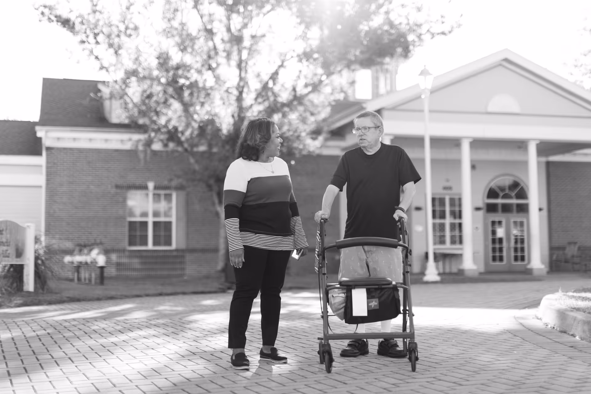 An elderly man using a walker is standing and talking with a woman outside a brick building with white columns and large windows. The scene is outdoors on a paved area with trees and sunlight in the background.