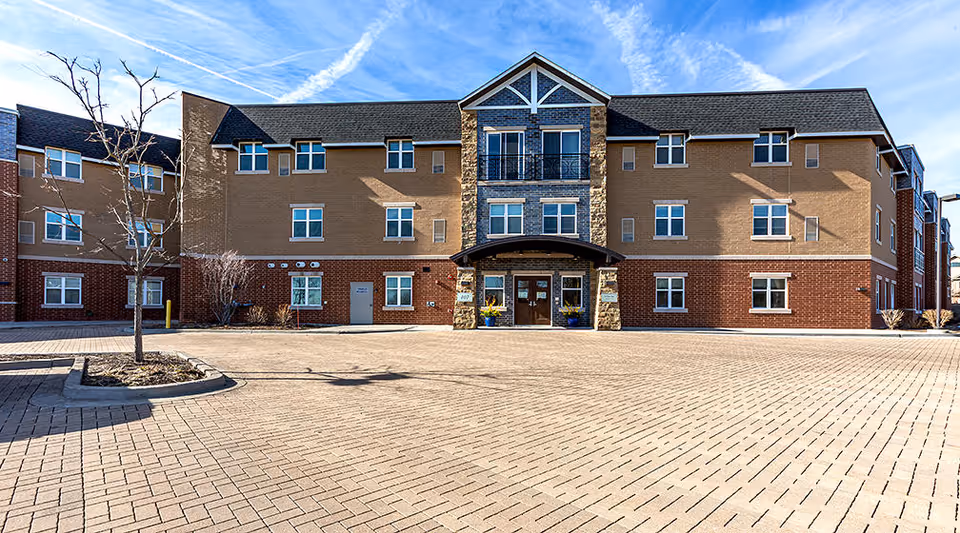 Front exterior view of a three-story senior living facility building with a brick and stone facade, multiple windows, a covered entrance with double doors, and a paved driveway in front. There are a few small trees and planters near the entrance under a clear blue sky.