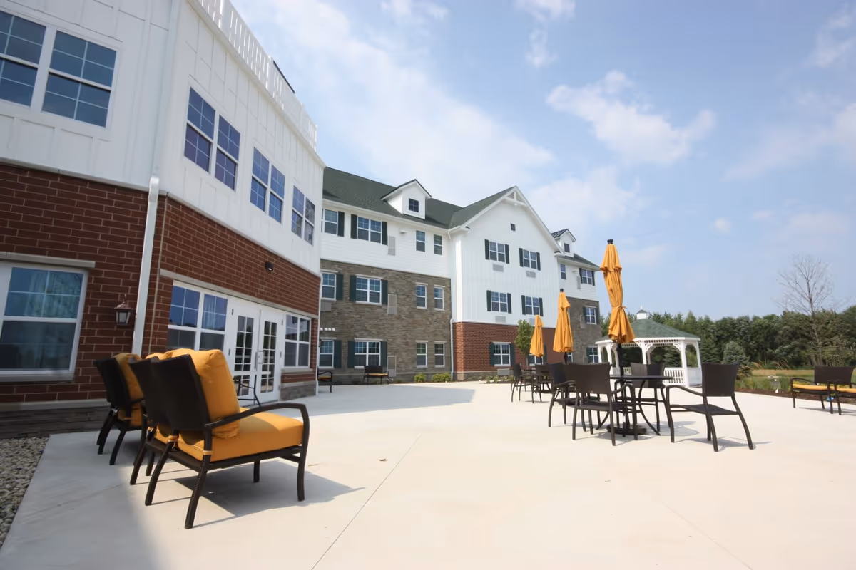 Outdoor patio area at Heritage Woods of Huntley featuring several black chairs with yellow cushions, black tables with closed yellow umbrellas, a white gazebo, and a multi-story building with white siding, brick, and green shutters under a partly cloudy sky.