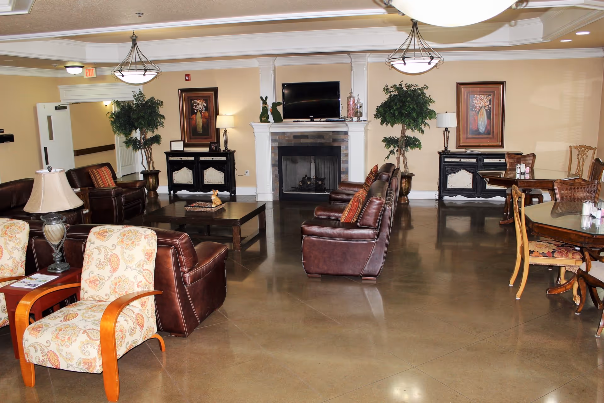 A spacious senior living common area with polished concrete floors, featuring multiple seating options including patterned armchairs and brown leather chairs arranged around a central coffee table. The room has a fireplace with a TV mounted above it, two black side cabinets with table lamps and framed artwork above them, and two large potted plants. There are also wooden dining tables with chairs on the right side of the room. The ceiling has recessed lighting and two hanging light fixtures.