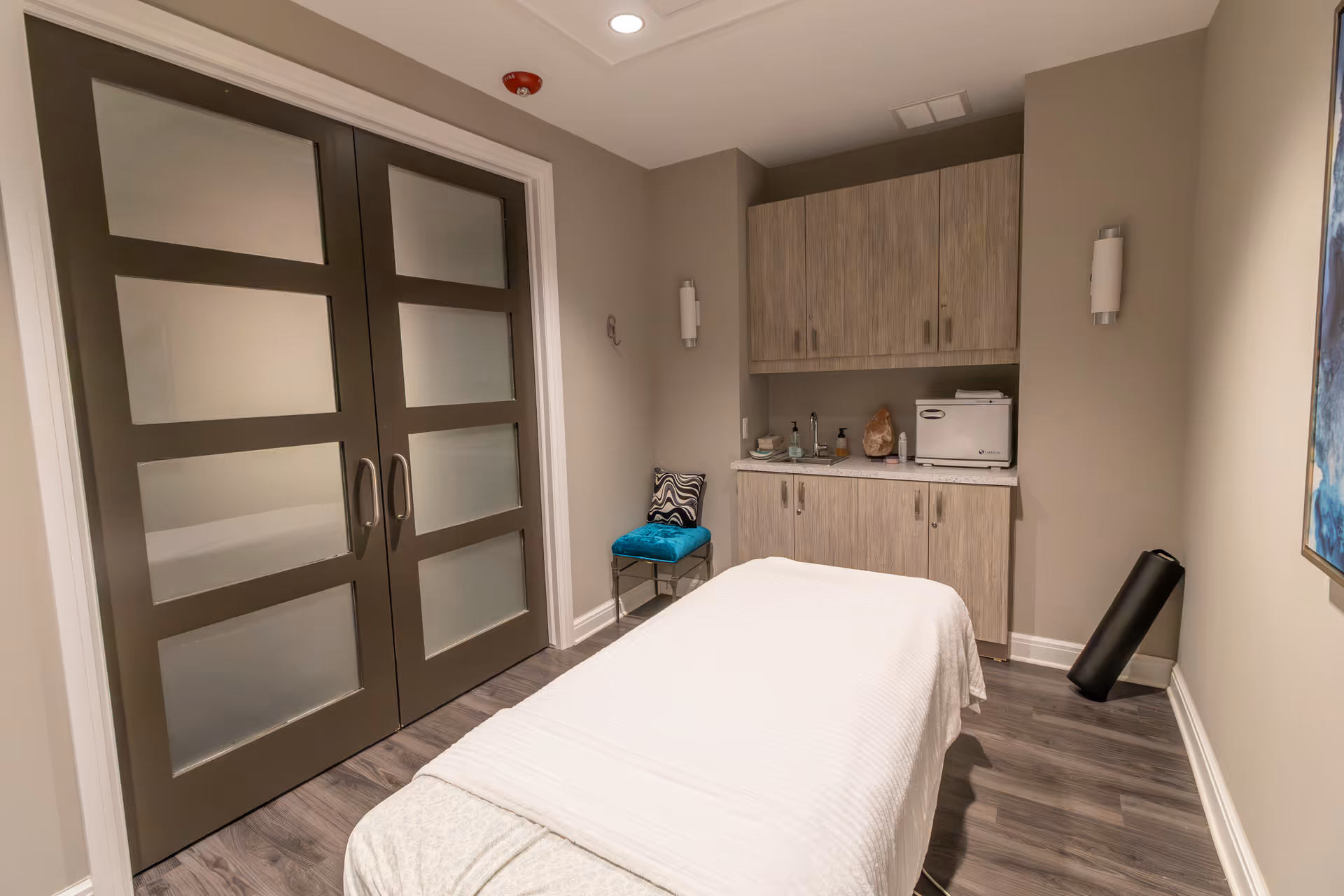 A calm treatment room with a massage table, frosted double doors, wood cabinets and a small teal chair.